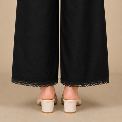 Black wide-leg trousers with a delicate black lace hem, shown from the back on a model wearing off-white block-heeled mule sandals against a solid beige background.