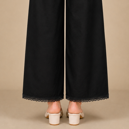 Black wide-leg trousers with a delicate black lace hem, shown from the back on a model wearing off-white block-heeled mule sandals against a solid beige background.